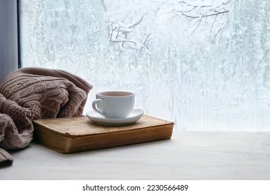 Winter background. tea cup, book, sweater and winter frozen window. cozy mood, home comfort in snowy cold weather. festive winter season. Christmas, New Year holidays. copy space - Powered by Shutterstock