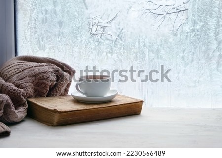 Winter background. tea cup, book, sweater and winter frozen window. cozy mood, home comfort in snowy cold weather. festive winter season. Christmas, New Year holidays. copy space