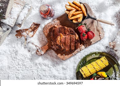 Winter BBQ with spicy marinated rib-eye steak and vegetables including corn and potato wedges served on rustic platters outdoors in snow