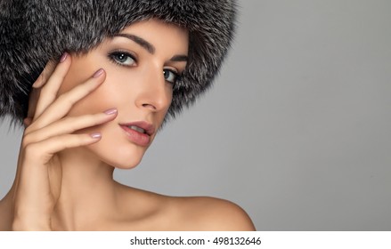 Winter beauty fashion. Beautiful face girl with trendy fur hat gesturing. Emotions. Professional makeup and manicure. High fashion portrait on grey background.  hand near the face