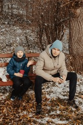 Winter Bonding: Brothers, Aged 8 And 17, Enjoying Tea On Snow-Covered Bench In Rural Park