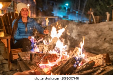 Winter campfire couple roasting marshmallows in firepit for smores at ski holiday resort. Asian happy woman relaxing after ski.