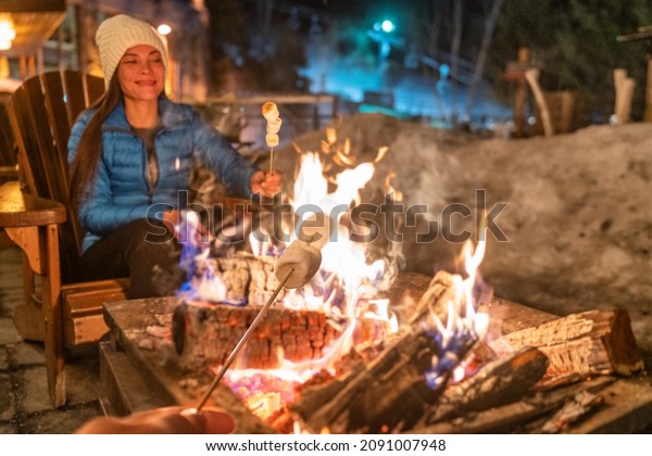 Winter campfire couple roasting marshmallows in\
firepit for smores at ski holiday resort. Asian happy woman\
relaxing after ski.