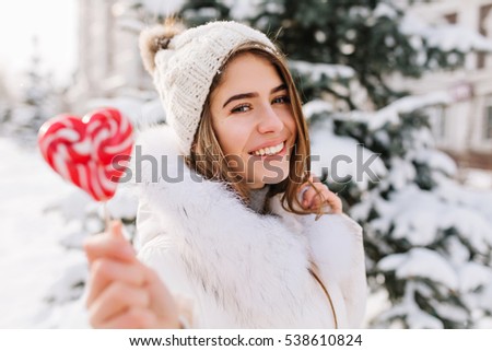 Winter closeup portrait charming joyful young woman in sunny winter morning with pink lollipop on street. White warm woolen knitted hat, enjoying cold weather, delicious. Happy time, positive emotions