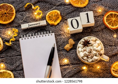 Winter composition. Wooden calendar January 1st Cup of cocoa with marshmallow, empty open notepad with pen, dried oranges, light garland on grey knitted background. Top view Flat lay Mockup - Powered by Shutterstock