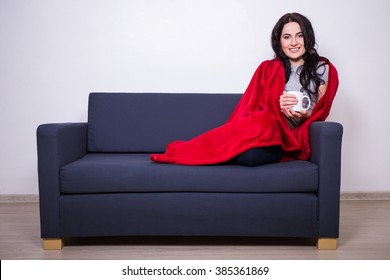 winter concept - young woman sitting on sofa with mug of tea wrapped in warm red blanket - Powered by Shutterstock