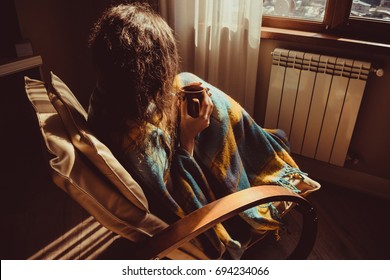 Winter Concept. Young Woman Sitting In Comfortable Modern Chair Near Radiator With Mug Of Tea Wrapped In Warm Plaid Blanket. Natural Light. Warm Atmosphere. Cup Hot Beverage. Heating Season. Cosy Home