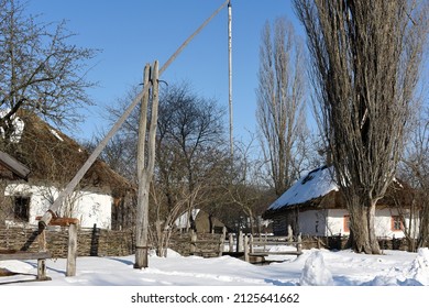 Winter countryside snowy landscape with snow-covered wooden well and old houses in the background Sunny day, bright blue sky. Fresh air. Green tourism, ecotourism in the countryside. Winter vacation