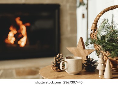 Winter cozy living room in farmhouse. Stylish cup of warm tea, basket with fir branches, wooden trees and star, pine cones against burning fireplace. Modern christmas rustic eco friendly decor - Powered by Shutterstock