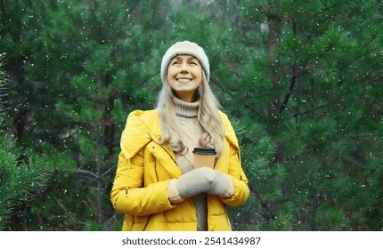 Winter day, happy smiling mature woman holding cup of coffee drink enjoys snow, warm weather against Christmas tree, wearing yellow jacket, hat, standing in snowy forest with snowflakes - Powered by Shutterstock