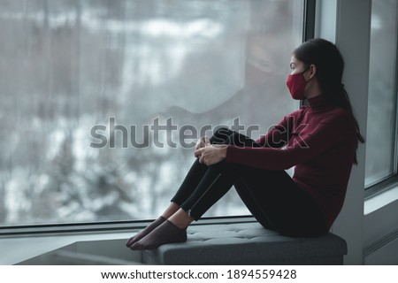 Winter depression because of coronavirus confinement. Sad Asian woman alone during city lockdown wearing face mask indoors at home covid. Anxiety, stress, mental health issues.