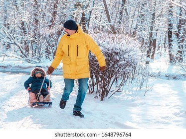 Winter Family Walk Outdoors: Photo Of Dad Running Along Snowy Road And Pulling Sled With Toddler Child Sitting In It. Father And Son Sledding And Having Fun Together. Focus On Child Face. Copy Space