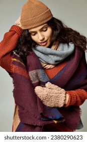 winter fashion, curly brunette woman in layered clothes with warm scarf and mittens on grey backdrop