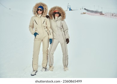 Winter fashion. Full-length portrait of two fashion model girls posing in white downy overalls against the backdrop of a snowy landscape. Alpine skiing, active winter recreation.