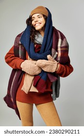 winter fashion, joyous model in layered clothing, warm hat and scarfs posing on grey backdrop