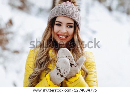 Winter fashion portrait of blonde student woman in knitted hat, gloves and yellow coat walking in snow park. Cold season.