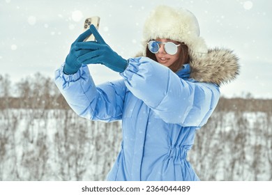 Winter fashion. Portrait of a fashionable girl posing in blue ski suit against a snowy winter landscape taking a selfie with smartphone. Sport in the winter mountains. 