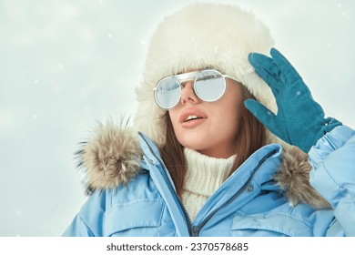 Winter fashion. Portrait of a fashionable girl posing in blue ski suit, white fur hat and stylish sunglasses  against a snowy winter landscape. Sport in the winter mountains. Copy space. - Powered by Shutterstock