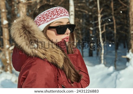Winter fashion. Sideview portrait of a beautiful fashion model girl posing in a red down overall and sunglasses in a winter snowy park. Winter holidays.