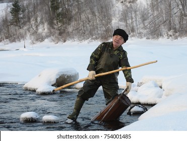 Winter Fishing On The Lamprey (Petromyzontiformes). A Man In Waders Is Knee-deep In The Water. He Drew A Fishing Trap From The Pond. The Trap For Catching Lamprey Is Heavy, There Is Prey In It.