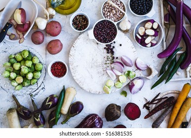 Winter Food Ingredients. Variety Eggplants, Colorful Carrots, Beetroot, Potatoes, Black And Red Beans, Brussels Sprout, Herbs And Spices On Chopping Board, Over Rustic White Table. Top View. 