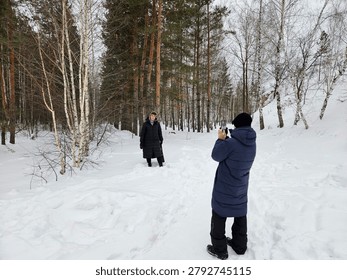 Winter forest snowy path. Two people stand on snow-covered ground. One person captures photo with camera. Trees surround them, mostly bare. Snow blankets the ground and branches