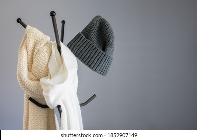 Winter Garment Accessory, Minimal Style Of White Cotton Scarf, Soft Knitted Scarf, And Casual Knitted Hat Hanging On A Coat Stand Against The Wall. Hanger For Clothes And Jacket, Simple Indoor Rack.