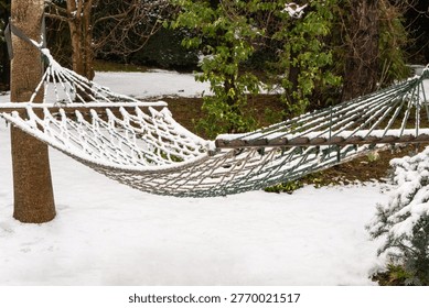 Winter hammock, stretched between two wooden posts amidst snowy landscape. Thick layer of freshly fallen snow blankets hammock's mesh fabric, giving it white appearance. Area is also covered in snow.