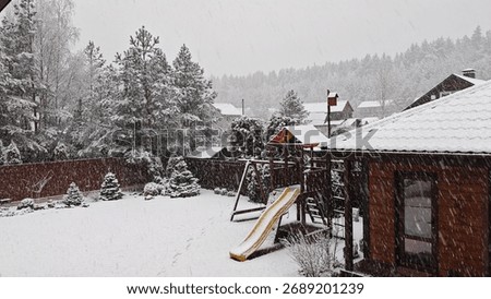 Winter has returned in the middle of spring. Heavy snow is falling with large snowflakes. Snow lies on the grassy lawn, playground, on the trees and on the roof of a wooden house