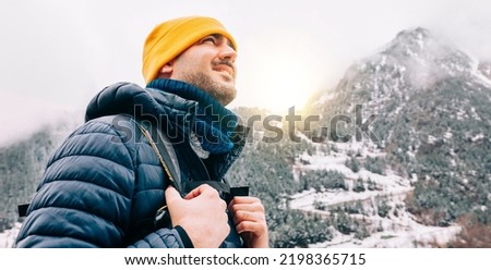 Winter hike on snow mountain young happy hiker man climbing. Europe travel adventure trek in nature landscape. Young cheerful person wearing yellow hat, blue jacket for cold weather and bag.