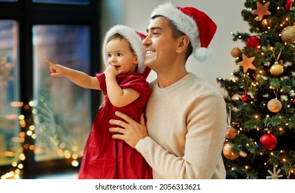 winter holidays and family concept - happy middle-aged father and baby daughter in santa hat looking through window over christmas tree at home - Powered by Shutterstock