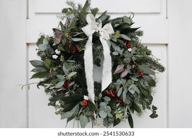 Winter holidays festive still live. Advent floral wreath. Green fir, juniperus, pine, eucalyptus tree branches and ivy. Red berries with silk ribbon. Blurred white wooden door background, home decor. - Powered by Shutterstock