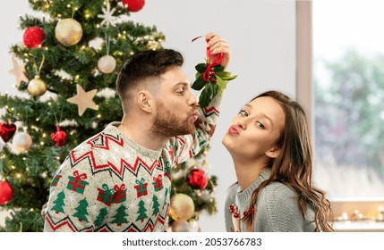 winter holidays, people and traditions concept - happy couple in ugly sweaters kissing under the mistletoe over christmas tree on background