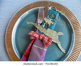 Winter holidays place setting with blue midcentury modern china on gold charger with festive napkin, ribbon and deco deer ornament. Blue rattan background.