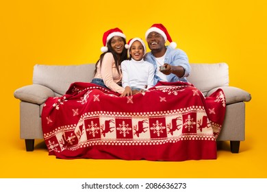 Winter Holidays. Portrait Of Cheerful Black Family In Red Santa Claus Hats Celebrating Xmas Evening And New Year At Home Sitting On Couch Under Cozy Warm Red Blanket Watching Tv Holding Remote Control