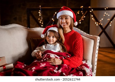 Winter Holidays. Portrait Of Cheerful Woman And Small Girl In Santa Claus Hats Celebrating Xmas Evening And New Year At Home, Sitting On The Couch Under Cozy Warm Red Blanket, Watching Tv Cartoons