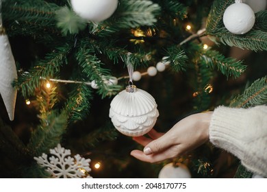 Winter holidays preparations.Hand in cozy sweater holding modern white bauble at christmas tree lights. Woman decorating stylish boho christmas tree with vintage ornament. Atmospheric hygge home