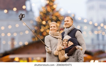 Winter Holidays, Technology And People Concept - Happy Family Taking Picture With Smartphone And Selfie Stick Over Christmas Market Background