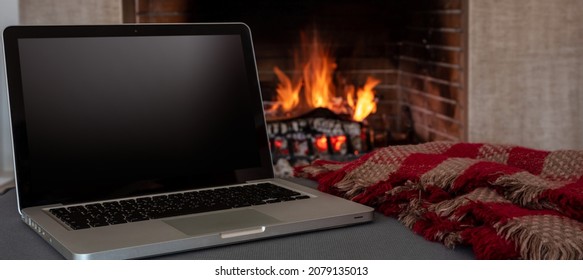 Winter Home Office Concept. Blank Screen Laptop On Burning Fireplace Background, Black Empty Computer Display, Cozy Room Interior. Christmas Warm House