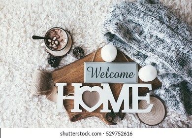 Winter homely scene, scandinavian style. Warm knit sweater, candles, cup of sweet cocoa with marshmallows and other decor on tray in bed. Wooden craft letters Welcome Home. Lazy cold weekend.