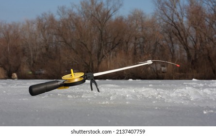 Winter Ice Fishing. Fishing Rod With Reel, For Ice Fishing With Float Equipment, Stands On Ice Near Hole. Side View. In Background Are Trees.