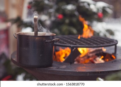 Winter Image Of Festive Decorated Christmas Street. Christmas Image Of  Iron Pan With Mulled Wine On Fire. Fir Trees Background. Cooking On Fire. Festive Drink. New Year Decoration