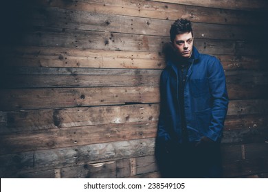 Winter Jeans Fashion Man With Short Dark Hair. Wearing Blue Jeans, Jacket And Brown Leather Gloves. Leaning Against Old Wooden Wall.