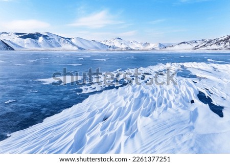 Winter landscape of frozen Baikal Lake in February cold sunny day. Сoastal mountains are covered with snow. On the surface of the ice, snow crusts in form of frozen waves. Beautiful winter background