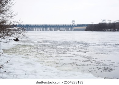 Winter landscape with a frozen river and a large dam in the background. Snow-covered trees and ice formations create a serene and cold atmosphere. - Powered by Shutterstock