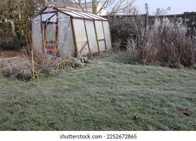 Winter landscape garden greenhouse the old glass wood abandoned building frozen in heavy white frost with misty broken opaque pane, icy lawn plants trees season frosty weather grey sky in England uk