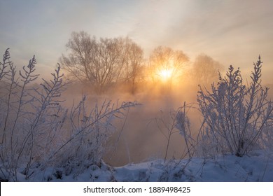 a winter landscape with a snow-covered river bank, trees covered with hoarfrost, fog and the rising sun over the river
