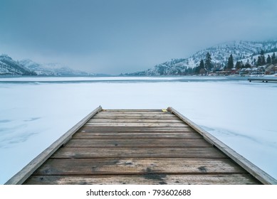 Winter Landscape Of Wooden Dock In Foreground On Frozen Lake With Snow Covered Mountains And Fog In Background