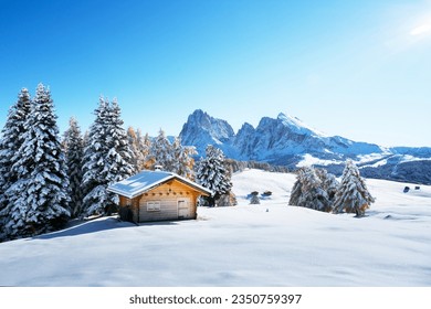 Winter landscape with wooden log cabin on meadow Alpe di Siusi on blue sky background on sunrise time. Dolomites, Italy. Snowy hills with orange larch and Sassolungo and Langkofel mountains group - Powered by Shutterstock