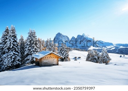 Winter landscape with wooden log cabin on meadow Alpe di Siusi on blue sky background on sunrise time. Dolomites, Italy. Snowy hills with orange larch and Sassolungo and Langkofel mountains group Stock fotó © 